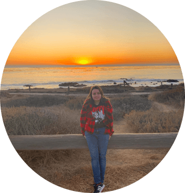J.J. Hale wearing a red shirt and black t-shirt standing in front of a wooden gate smiling, the ocean is in the background with a beautiful sunset.
