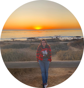 J.J. Hale wearing a red shirt and black t-shirt standing in front of a wooden gate smiling, the ocean is in the background with a beautiful sunset.